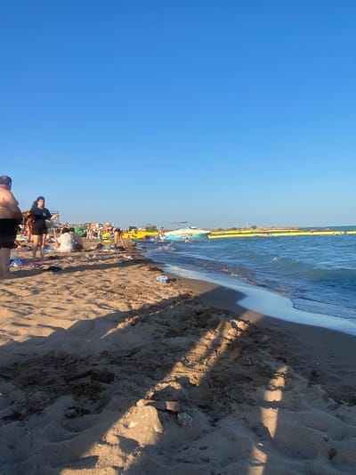 The image shows a sandy beach scene at the city of Shybartubek, Kazakhstan. The sky is clear and blue. Several people are scattered on the beach, some sitting and relaxing on towels, others standing and interacting. In the foreground, shadows from unseen objects are cast across the sand. Further back, towards the water, there are more people, some in the water, and several yellow buoys extending into the distance. A white boat is also visible on the water. The overall impression is a lively summer day at the beach.