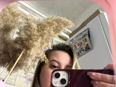 A woman is taking a selfie in a mirror. She is holding a phone with a dark red or burgundy case. In the background, there is what appears to be a refrigerator with magnets or photographs attached. There's also a decorative plant with fluffy, wheat-like plumes. The location information (Port-Katon, Russia) is not discernible from the image itself.