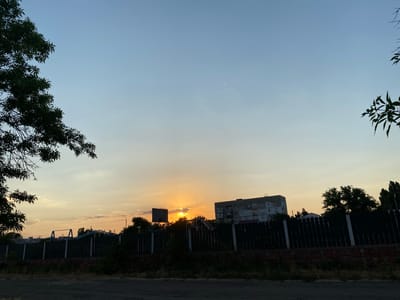 The image depicts a sunset over a residential area in Voznesensk, Ukraine. The sky is a gradient of pale blue at the top transitioning to warm hues of yellow and orange near the horizon, with scattered clouds. In the foreground, a dark, dirt road is visible. Behind the road, a dark wooden fence runs horizontally across the frame, with a brick foundation at its base. Beyond the fence, glimpses of trees, playground equipment, and a multi-story building are visible. The setting sun is partially obscured by clouds and appears as a bright, orange orb. Silhouettes of trees and a basketball hoop are also present. The overall scene captures a quiet, urban evening.