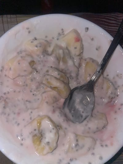 A close-up, high-angle shot captures a white bowl filled with a creamy breakfast or snack item, likely yogurt or a similar dairy-based mixture. The main ingredients visible are numerous slices of ripe banana and a generous amount of small, black, speckled chia seeds, which are dispersed throughout the creamy base. Hints of a light pinkish hue can be observed in various parts of the mixture, suggesting the presence of berries or fruit purée. A silver spoon is resting within the bowl, partially submerged in the food, indicating that the meal is either being consumed or is ready for consumption. The lighting is soft, casting a slight sheen on the surface of the creamy mixture and the spoon. The background immediately surrounding the bowl is dark, with a subtle vertical striped pattern barely discernible on the left, possibly a placemat or tablecloth. No people are visible, and there is no discernible text. The image provides no specific cues regarding the time of day, weather, or location within Chirpan, Bulgaria.
