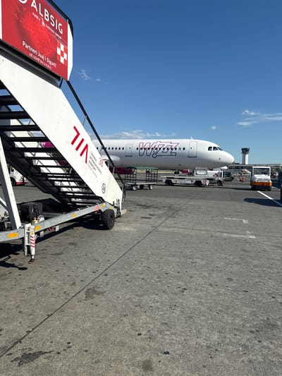The image shows an airplane parked on an airport tarmac, with stairs leading up to the aircraft. The airplane is white with the Wizz Air logo on its side. The stairs are white with red lettering that reads "TIRANA INTERNATIONAL AIRPORT NENE TEREZA". There are also several airport vehicles visible, including a tug and a baggage cart. The sky is blue with some clouds. There are no people clearly visible in the image. The scene is set at an airport, likely Tirana International Airport "Nene Tereza" in Albania, given the text on the stairs. The image does not provide any specific information about the city of Kruja.