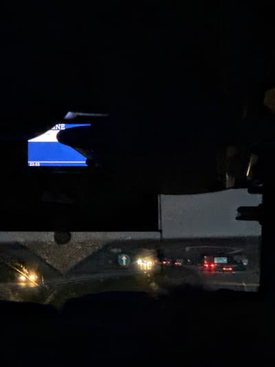The image depicts a night scene, likely from inside a vehicle. In the foreground, a dark silhouette of a hand is visible, obscuring a portion of a digital display screen. This screen shows the word "NE" in blue text, with a white horizontal line below it. At the bottom of the screen, the time "22:55" is displayed.
The mid-ground reveals a wet road surface with multiple vehicles. Headlights and taillights of cars are illuminated, indicating traffic. A blue circular sign with a white arrow pointing upwards is visible on the right side of the road. The overall environment suggests a highway or a busy road in a city.
Although the specific location of Medemciems, Latvia, is mentioned, there are no distinct visual cues within the image that definitively confirm this location. The image is characterized by low light conditions, reflections on the windshield, and the presence of artificial lights from vehicles and signage.