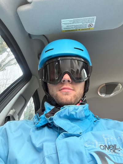 A male individual is depicted in a close-up, seated inside a vehicle. He wears a light blue ski helmet with dark vents and black ski goggles covering his eyes and upper nose. A light blue ski jacket with a high collar and an "O'NEILL" logo with a graphic design on the right chest/sleeve area is visible. A dark undergarment is present around his neck. The vehicle's interior features a light grey headliner and sun visor, with a yellow "WARNING / AVVERTENZA" label affixed to the visor. A ceiling light fixture is partially visible on the right. Through the vehicle's window on the left, bare tree branches are visible against an overcast sky. The specific location within the city of Kolašin, Montenegro, cannot be determined from the image.