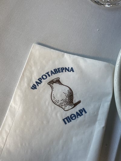 The image shows a close-up, overhead view of a white paper napkin with blue Greek lettering and a brown drawing of a jug. The napkin is placed on a textured, light gray surface, likely a tablecloth. The writing on the napkin reads "ΨΑΡΟΤΑΒΕΡΝΑ" and "ΠΙΘΑΡΙ". The jug drawing is in the center, below the first line of text and above the second. The text and drawing are printed on the napkin. To the right of the napkin, a sliver of a white, curved object, possibly a plate or bowl, is visible. The overall scene suggests a table setting at a restaurant. The location of Ofrynio Beach, Greece, is inferred to be the setting for this image, suggesting the restaurant is likely a seafood taverna ("Ψαροταβέρνα") located there. There are no people or actions depicted in the image, only static objects.