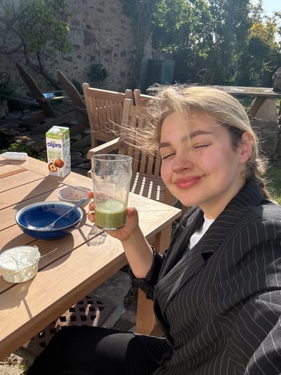 A young woman sits at a wooden table in an outdoor setting. She is wearing a black and white striped blazer, black pants, and has long blonde hair. Her left hand is holding a glass of a green smoothie. The table is covered in a wooden tabletop and has a carton of Alpro hazelnut milk, a blue bowl with a silver fork, a small bowl of a white substance, and a small bowl containing a red substance on it. The woman is smiling at the camera, and her eyes are closed. The location is a patio in Crail, Scotland. The environment is surrounded by trees and plants, and there is a stone wall in the background. The patio is surrounded by wooden chairs. The sun is shining brightly.