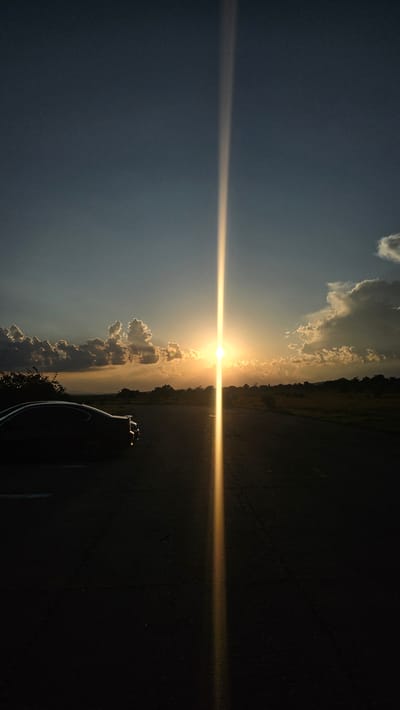 The image captures an outdoor scene at sunset in Buhovtsi, Bulgaria. The sky exhibits a gradient from dark blue at the zenith to orange and yellow hues near the horizon, where the sun is centered. Various cumulus clouds are scattered across the sky, with some illuminated by the setting sun and others in shadow. A prominent vertical light streak, originating from the sun, extends upwards into the sky and downwards onto the dark asphalt road surface. A dark-colored vehicle, discernible as a coupe or sedan silhouette, is parked on the left side of the road, facing away from the viewer. The background landscape consists of silhouetted trees and fields extending towards the horizon. No individuals are present in the scene.