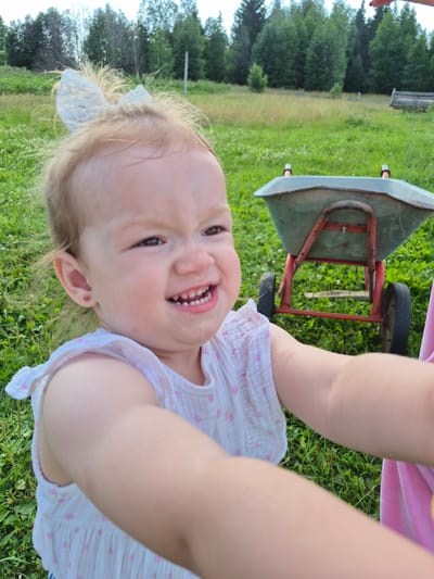 A close-up shot shows a young Caucasian girl with blonde hair and a white bow on her head, wearing a white dress with pink polka dots. She has a playful expression, with her mouth open in a smile and her eyes squinting. Her left earlobe has a small pink stud earring. The background is a grassy field with trees and a red and gray wheelbarrow. The image appears to have been taken outdoors in a rural setting, possibly in the city of Дойная, Russia.