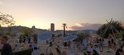 This is a wide-angle, panoramic photograph taken at sunset at the Summer-Voest beach club, a popular summer destination located on the banks of the Danube River in Linz, Austria. The scene depicts a lively atmosphere with numerous people relaxing on lounge chairs on a sandy beach.
In the foreground, several individuals are visible, some seated and others standing. A woman with blonde hair pulled back in a bun is in the left foreground, wearing a dark top. Behind her, a man in a white shirt is visible, seemingly yawning. Other people are scattered throughout the beach area, enjoying the evening.
The middle ground features a collection of beach umbrellas, some blue with the Zurich logo and others yellow, providing shade. Several people are gathered around these umbrellas, some standing and conversing, others seated. A tall palm tree stands prominently to the right of the blue umbrellas. Further back, a distinctive modern skyscraper with a curved facade dominates the city skyline, a landmark of Linz. Other buildings of varying heights and architectural styles are also visible, forming the urban backdrop.
In the background, the Danube River is hinted at by the presence of a boat or barge. The sky is filled with the warm hues of sunset, transitioning from pale orange to soft pink and purple, with some wispy clouds. A conical, teepee-like wooden structure with lights is visible on the right side of the image, adding to the festive ambiance.
The overall impression is one of a vibrant social gathering at a riverside beach club, with a blend of urban scenery and natural elements under the soft light of dusk.