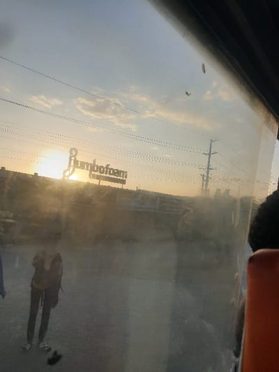 The image depicts a view through a car window, reflecting the outside scenery. The sunset creates a warm glow behind a Jumbofoam sign. Utility poles line the horizon. A person's reflection appears in the lower right corner, along with a glimpse of a blue object. The overall image is somewhat blurry and unclear due to the reflections on the window.