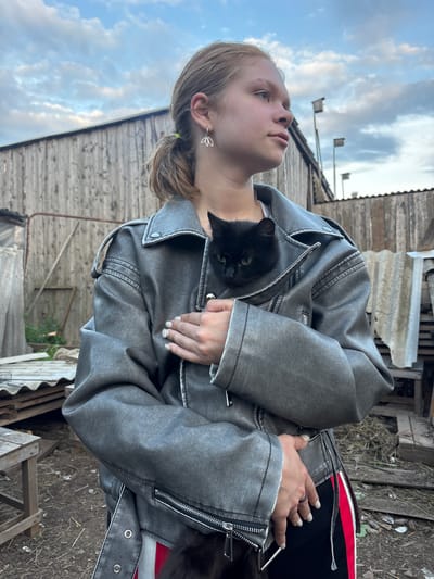 The image features a young woman with blonde hair, wearing a grey denim jacket, holding a black cat. The cat is nestled in the crook of her arm, with its face peeking out from the jacket. The woman is looking upwards with a thoughtful expression. Her left hand, adorned with a ring and white nail polish, rests on her chest. She is also wearing a delicate leaf-shaped earring.

The setting appears to be outdoors, with a wooden structure and weathered materials visible in the background, suggesting a rural or rustic environment. The sky is partly cloudy, indicating it might be daytime with mixed weather conditions. The overall mood of the image is serene and intimate, highlighting the bond between the woman and her pet. The orientation of the image is vertical.