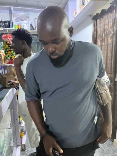 A bald man with a beard is standing in what appears to be a store. He is wearing a grey shirt and black pants, holding a black object in his right hand. Behind him, another person is visible, also appearing to be in the store. Shelves stocked with various items are evident in the background, along with decorations. The location is likely in Dushepe, Nigeria.