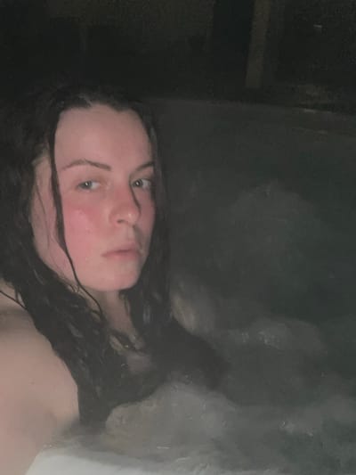 A person with wet, dark curly hair and flushed skin looks at the camera. Her face is centered in the lower half of the image. The background is dark, with unclear shapes in the upper right suggesting water or steam. Part of a dark wall is visible on the left. The lighting is dim, giving the image a low-contrast, somewhat blurry quality.