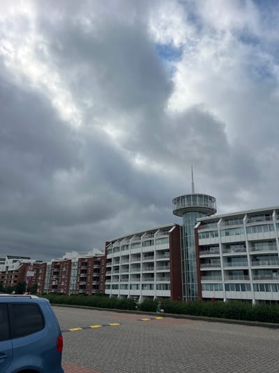 A portrait-oriented, downward-angled shot shows a modern building complex in Wilhelmshaven, Germany, under a heavily clouded sky. The building on the right is white with multiple balconies and glass-fronted windows. A distinctive circular structure with a spire juts out from a connecting section. Below it, a series of red brick buildings with white-trimmed windows continues the architectural line. In the foreground, a blue car is parked on a grey, patterned paved surface. A low green hedge runs along the base of the buildings, separating them from the pavement. Two figures are visible near the hedge: one in a green shirt appears to be interacting with a white object, possibly a ball, while another individual in a red garment stands further back. The weather is overcast, with dramatic, dark grey clouds dominating the sky, interspersed with lighter, brighter patches, suggesting a potentially stormy or unsettled atmosphere. The time of day is not explicitly discernible due to the weather conditions, but the general lighting suggests daytime. The scene is one of a modern urban or residential environment, with a focus on the contemporary architecture and the interplay of clouds and structures.