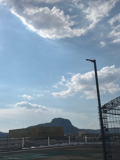 The image depicts a view of the Nestlé factory in Singen (Hohentwiel), Germany. In the foreground, a dark lamppost stands to the right, silhouetted against a blue sky with scattered white clouds. A metal cage structure is also visible to the far right. In the middle ground, a long, yellow building with orange stripes is prominent, bearing the "Maggi" and "Nestle" logos. Rows of arched structures, possibly covered parking or storage, are in front of the factory. In the background, a rounded, tree-covered hill, likely the Hohentwiel, rises above the factory and other buildings, including a water tower. The overall scene is an outdoor view of an industrial area with a natural landscape element.