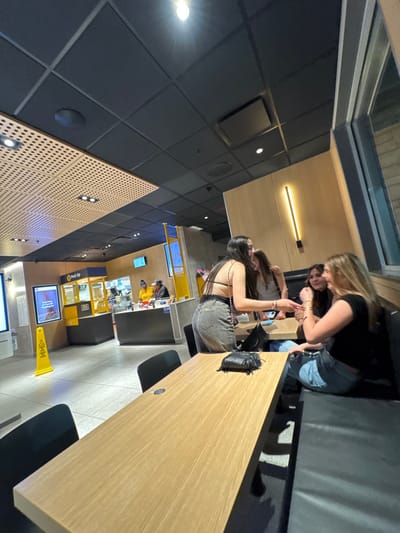 The image depicts the interior of a McDonald's restaurant, likely located in South Bend, United States.
**Environment:** The space features a modern design with a dark, segmented ceiling containing recessed circular and linear lighting. A section of the ceiling on the left has a light-colored, perforated acoustic panel. Walls are primarily clad in light wood paneling, with some darker stone or concrete elements visible near a large window on the far right. The flooring consists of light-colored tiles in a grid pattern. In the mid-ground, a long, light wooden communal table with dark chairs on the left is present. A dark, upholstered booth runs along the right wall. Artificial lighting illuminates the entire scene, suggesting it is evening or night. In the background, a "Pick Up" counter in yellow and black is visible, along with additional service counters and digital displays. A yellow "wet floor" warning sign stands near the service area.
**People and Interactions:**
Four young females are seated at the booth and table on the right side of the image.
1. **Female 1 (standing):** Positioned partially behind the wooden table, facing towards the booth. She has long dark hair, wears a black backless top with thin straps, and light-colored denim jeans. She is holding a dark-colored smartphone in her left hand, extending it forward, possibly for viewing by the others. She wears bracelets on her left wrist.
2. **Female 2 (seated):** Partially obscured, she is positioned at the booth, looking towards Female 1. She has long dark hair and wears a light grey top.
3. **Female 3 (seated):** Positioned at the booth to the left of Female 4. She has dark hair, wears a dark top, and is smiling while looking towards Female 4, with her left hand near her mouth.
4. **Female 4 (seated):** Positioned at the booth on the far right. She has long blonde hair, wears a black top and light blue denim jeans. She is smiling and looking towards Female 3, with her right forearm resting on the table.
A black fringed handbag rests on the wooden table in the immediate foreground.
In the background, behind the service counters, two individuals, presumably McDonald's staff, are visible. One wears a yellow uniform top and a dark baseball cap; the other wears a dark uniform top and a baseball cap. They appear to be engaged in work-related tasks behind the counters.