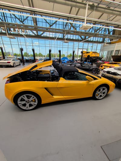 The image shows a bright yellow Lamborghini with its hood open, parked indoors in a showroom or garage. The car's engine is visible. The setting appears to be a modern building with large windows and a high ceiling, with a steel support structure overhead. Sunlight streams in through the windows, suggesting it is daytime. Outside the windows, trees and other cars can be seen, indicating a possibly urban or suburban environment. Another white car is parked behind the yellow Lamborghini. Several other vehicles, mostly sports cars, are visible in the background, lined up in the showroom. The image is oriented vertically. There is no visible text in the image. The scene is clean and well-lit, suggesting a professional setting like a car dealership or a private collection.