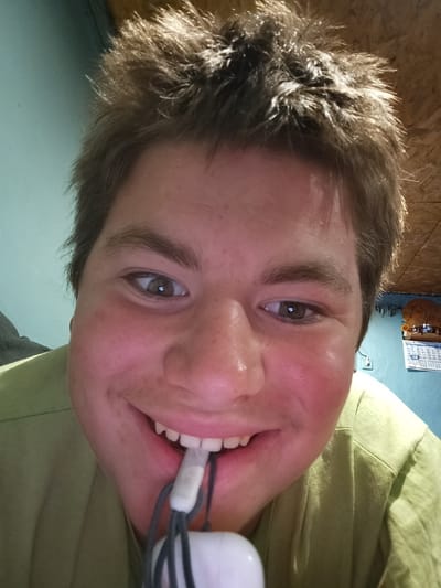 A close-up selfie of a person with short, spiky brown hair and light skin is presented. The person is smiling, revealing their teeth, and has a pair of white earphones with gray cords held in their mouth. They are wearing a light green shirt. The background is a pale turquoise wall with a partially visible calendar hanging on it. The lighting appears to be indoor. There's no definitive information to confirm the location as Chernik, Bulgaria.
