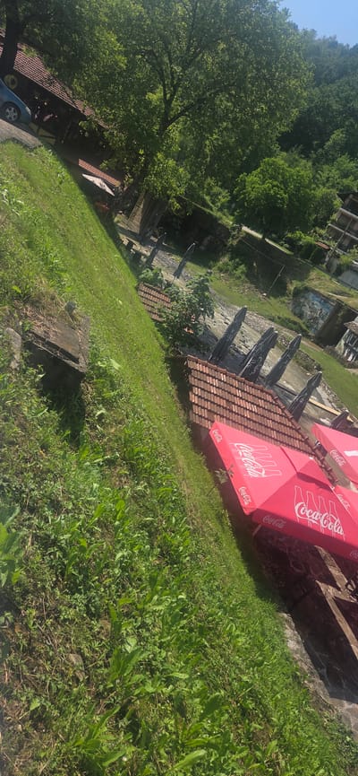 The image, taken with a tilted perspective, depicts an outdoor scene in Beli Osam, Bulgaria, under clear, sunny conditions. In the lower right, a series of red umbrellas with the Coca-Cola logo are arranged, indicating an outdoor seating area. Adjacent to these umbrellas is a building section with a reddish-brown tiled roof. To the left of the umbrellas, a steep, grassy embankment dominates the lower foreground, showing varied green vegetation.
Further into the scene, behind the initial tiled roof, is a flowing body of water, likely a river or stream. Along its banks, additional outdoor umbrellas, appearing dark in color, are visible. On the opposite side of the water, a paved road runs, and beyond it, a structure with a painted mural is discernible on a retaining wall. Another multi-story building with balconies is situated further up on the far bank.
In the upper left portion of the image, a light blue car is parked on a surface that appears to be part of a wooden deck or paved area, adjacent to a larger building. This building features a reddish-brown tiled roof, similar to the one in the foreground, and light-colored walls. Abundant green foliage, consisting of numerous trees with dense canopies, frames the buildings and lines the surrounding hillsides. The background is dominated by heavily forested hills rising under a clear blue sky. No explicit human figures or direct interactions are clearly visible.