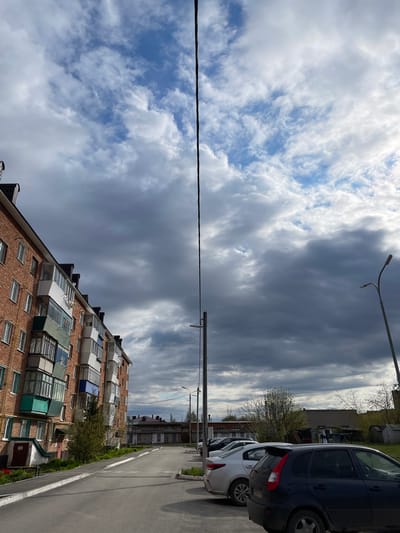 The image shows a street in Urussu, Russia, with an apartment building on the left, cars parked on the right, and a cloudy sky overhead. The apartment building is made of red brick and has multiple balconies of various colors and designs. A utility pole with a power line spans across the top half of the image. A street lamp is visible above the parked cars. The road appears to be asphalt. No people are visible in the shot. The weather is overcast.