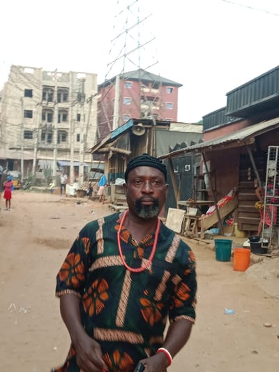 A man in traditional attire stands in front of a dusty street lined with small, ramshackle buildings. He wears a dark green shirt with orange floral patterns and a red beaded necklace. In the background, an unfinished concrete building and a red apartment complex are visible, connected by a tall metal structure. The scene suggests an urban setting with a mix of modern and informal architecture. Other people are distantly present in the background.