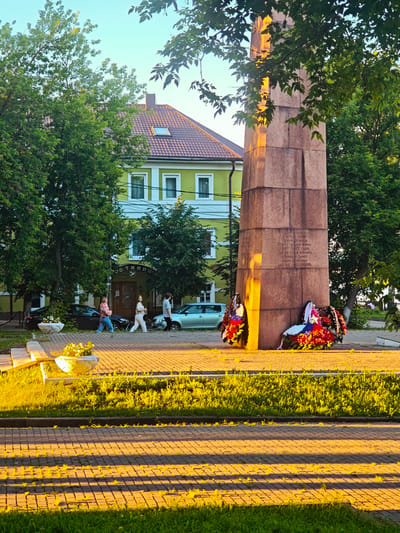 This image captures a solemn monument in Kimry, Russia, bathed in the warm, golden light of late afternoon or early evening. The monument, a tall, reddish-brown stone obelisk, is adorned with two large, colorful wreaths at its base, featuring red, white, and blue ribbons, suggesting a memorial for fallen soldiers or a national holiday observance.
Inscriptions in Russian are visible on the monument, with some text clearly decipherable: "МОСТИНИЦ", "ГЕРОЯМ ВЕЛИКОЙ ОТЕЧЕСТВЕНОй войны 1941-1945 годов", and parts of phrases about remembrance and freedom.
Behind the monument, a bright green building with a red tiled roof and white-trimmed windows stands, partially obscured by lush green trees. Three people are walking on a paved walkway in front of the building. The foreground features a brick-paved area with long, dramatic shadows cast by an unseen object, indicating the sun's low angle. Grass and decorative planters with flowers are also visible. The sky above is a clear, pale blue. The overall atmosphere is one of quiet reflection and remembrance, enhanced by the peaceful setting and the gentle, warm lighting.