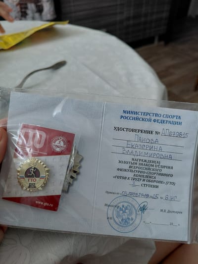 This media file shows a close-up of a person holding a plastic-encased document and a medal. The document appears to be an official certificate or award, with visible text in Russian indicating it is from the "MINISTRY OF SPORT OF THE RUSSIAN FEDERATION". The certificate is numbered "ΑΠ679615" and contains handwritten names: "ПАНОВА ЕКАТЕРИНА ВЛАДИМИРОВНА". It also mentions being awarded a "GOLDEN BADGE OF DISTINCTION" for the "ALL-RUSSIAN PHYSICAL CULTURE AND SPORTS COMPLEX 'READY FOR LABOR AND DEFENSE' (GTO)". The date on the certificate is partially visible as "03 FEBRUARY 2025". A blue stamp with the Russian coat of arms and the text "MINISTRY OF SPORT OF RUSSIA" is also present.
The medal, also enclosed in plastic, is gold-colored and features the "GTO" logo with the number "10" on it, suggesting a specific achievement level. A small red card with the website "www.gto.ru" is visible next to the medal. The background is a soft, light-colored fabric, possibly clothing or bedding, and a dark, checkered pattern is visible in the upper left corner. The lighting suggests an indoor setting, likely during the day, with no strong indicators of weather or specific time of day beyond the date on the document. The focus is on the documents and the medal, implying an event related to sports achievement or award.
