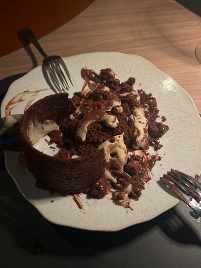 A close-up, overhead view presents a partially consumed dessert on a white, textured plate with an irregular, wavy rim. The plate rests on a light-colored wooden table surface, with a dark, possibly black, placemat positioned beneath it. The dessert consists of dark brown, crumbled cake or brownie material, mixed with a creamy, lighter-colored substance, likely ice cream or a similar dairy component, and liberally coated with dark brown chocolate sauce. Small, light-colored inclusions, consistent with nuts (possibly hazelnuts), are embedded within the dessert mixture. A significant, curved section of the original dark brown cake structure remains on the left side of the plate, partially upright. Two silver-colored dining forks are visible: one lies on the upper left edge of the plate, its tines pointed towards the dessert, while the second is partially visible on the lower right, resting off the plate. Both forks exhibit residue from the dessert. A single dark brown crumb is present on the dark placemat to the lower left of the plate. The scene is illuminated by warm, artificial light, suggesting an indoor setting within a restaurant or cafe located in Arles, France. An orange-brown wall or partition is visible in the upper left background, and the rim of a clear glass is faintly discernible in the upper right. No individuals are present in the image.