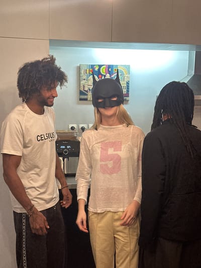 The image shows three people standing in what appears to be a kitchen. The man on the bottom is wearing a white "Celsius Live Fit" t-shirt and dark pants. He has curly hair and is looking at the person above him. The person in the middle is wearing a black Batman mask and a white long-sleeve shirt with a pink number 5 on it, paired with tan pants. Above them is a person in all black clothing with long dreadlocks. Behind them is an espresso machine, and a colorful abstract piece of art is visible on the wall. The location is Clichy, France.