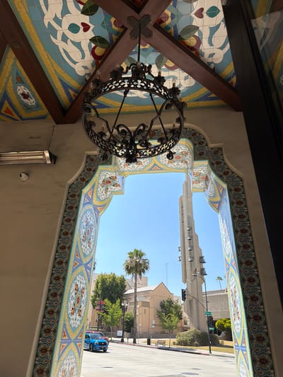The image shows an ornate arched entryway decorated with colorful mosaic tiles. Through the arch, a view of a street in Pasadena, California, is visible. A blue SUV is driving on the street. In the background, a tall concrete bell tower and a building with a steep roof are present. The sky is clear and blue. A black wrought-iron chandelier hangs from the ceiling inside the entryway. A security camera is mounted on the left wall.