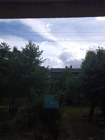 The image captures a view from a window, looking out onto a courtyard with trees and a building. The sky is partly cloudy with a bright, streaky cloud formation. Overhead, three power lines are visible. In the foreground, the dark interior of a room is implied by the upper edge of the frame, which appears to be a window frame or overhang.
Dominating the mid-ground are lush green trees with dense foliage, obscuring much of the view behind them. To the right of the trees, a building with a tiled roof and chimneys is partially visible. The building appears to be a multi-story structure, possibly residential. A small, bright blue rectangular object is situated amongst the trees on the left side of the image.
The ground in the courtyard is covered in grass and a dirt path. The overall impression is a somewhat overgrown, residential area. The scene is set in the city of Shakhtarsk, Ukraine.