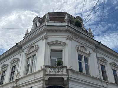 The image shows a corner of a white, multi-story building with ornate architectural details, located in Kolomyia, Ukraine. The building features multiple windows, some with decorative pediments above them, and a small balcony with a balustrade. The upper part of the building has a domed roof section with visible tiling and some greenery growing on the rooftop. A complex network of electrical wires crisscrosses the sky above the building. The sky is partly cloudy with patches of blue sky visible. The paint on the building shows signs of wear and peeling in places. The photograph is taken from a low angle, looking upwards at the building.