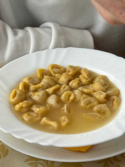 A close-up, high-angle shot depicts a white fluted ceramic bowl containing numerous tortellini submerged in a clear, light amber-colored broth, centered on a dining surface. The bowl rests upon a white underplate, with a folded yellow napkin positioned between the two plates. The table surface beneath is covered by a patterned tablecloth featuring golden and white hues. In the upper background, the chest and right arm of an individual, dressed in a light-colored, possibly white, textured garment, are partially visible. The specific location within Anzola dell'Emilia, Italy, cannot be determined from the visual evidence.