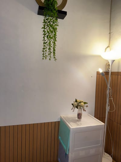A floor lamp with a white shade and a smaller adjustable light is positioned to the right of the image. The lamp is near a wooden-paneled wall. In front of the wall, a white plastic storage container is visible. On top of the container, a vase with a bouquet of white flowers sits. A trailing green plant is mounted on the wall above and to the left of the container. The lower portion of the wall features vertical wooden paneling. The scene is indoors, with a white wall and warm lighting.