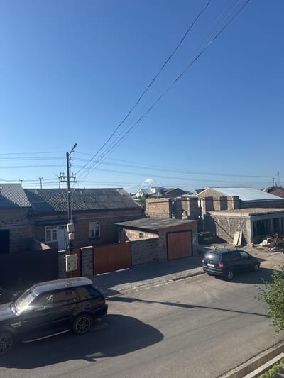 In Armavir, Armenia, under a clear blue sky, a sun-drenched street scene unfolds. On the left, a black Range Rover is parked, its sleek surface reflecting the bright sunlight. Across the asphalt road, a dark blue minivan is also parked. The street is lined with houses constructed from brick and concrete blocks, some appearing completed with corrugated metal roofs, while others are in various stages of construction. A wooden fence with a gate separates the street from one of the properties. Utility poles with visible wires and electrical boxes stand tall along the street. In the background, a snow-capped mountain, possibly Mount Ararat, is faintly visible against the horizon. The overall scene depicts a residential area in Armavir on a sunny day.