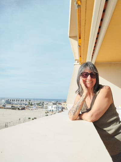 A woman with gray hair and sunglasses stands on a balcony in Palavas-les-Flots, France, with her left hand resting on her cheek, smiling. She has a tattoo on her left arm and wears a sleeveless olive-green shirt. The balcony overlooks a beach with palm trees and a marina in the background, with boats docked in the water. The sky is light blue. The building's facade is a light yellow hue.