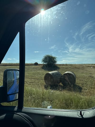 The image depicts a view from inside a vehicle, looking out at a rural landscape. The scene is dominated by a field of dry grass, likely hay, dotted with several large, round bales of hay. Two bales are prominently visible in the foreground, while another is further back in the field. A single, sparse tree stands in the middle ground, and a distant line of trees marks the horizon. The sky is a clear, bright blue with wispy white clouds, and the sun is shining brightly, causing lens flare in the upper left of the image. The presence of a side-view mirror reflecting a blue vehicle suggests the photo was taken while the vehicle was in motion or parked on a road adjacent to the field. The time of day appears to be late morning or early afternoon, given the angle of the sun and the brightness of the light. The overall impression is one of a rural, agricultural setting under a clear, sunny sky. No people are visible, and there is no discernible text in the image.