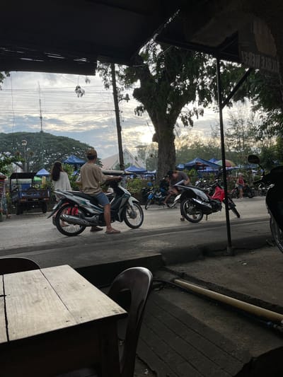 This image depicts a street scene, likely in Valencia, Philippines, featuring several motorcycles, people, and market stalls. In the foreground, a wooden table and chairs are visible, suggesting an outdoor seating area. A man wearing a tan t-shirt and blue shorts is riding a white and teal motorcycle, while a woman with long, dark hair stands beside him. Further down the street, other individuals are seen interacting with motorcycles, and market stalls with blue and purple umbrellas are set up. A small blue utility vehicle is also present, parked to the left of the street. The background shows trees, a church-like structure, and utility poles with numerous wires, indicating a typical urban or semi-urban environment. The lighting suggests it is either early morning or late afternoon, with the sun casting long shadows.