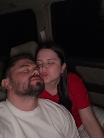 A couple is asleep, presumably in a vehicle. A woman with long, dark hair and a red shirt has her eyes closed. Her lips are puckered, perhaps in a kiss. A bearded man is nestled beneath her, wearing a white shirt. The scene is dimly lit, with the dark interior of the car visible in the background. They both appear relaxed and comfortable.