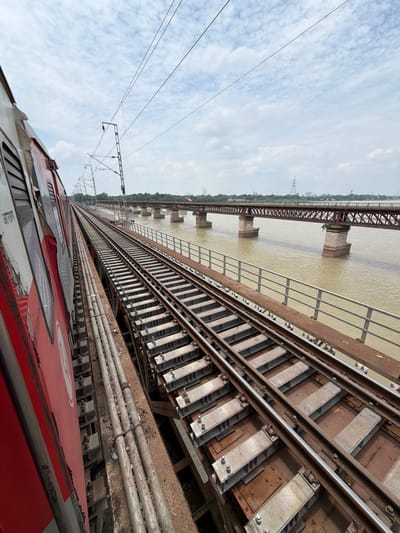 The image captures a view from a moving train on a bridge crossing a wide, murky river in Shuklaganj, India. The train, painted red and white, is on the left side of the frame, partially obscuring the view. The railway tracks stretch forward, disappearing into the distance. Parallel to the tracks is a metal railing. The river flows beneath the bridge, with a rusty, multi-span railway bridge visible in the background, also crossing the river. The sky is overcast with scattered clouds. The scene depicts a transportation infrastructure scene over a natural body of water.