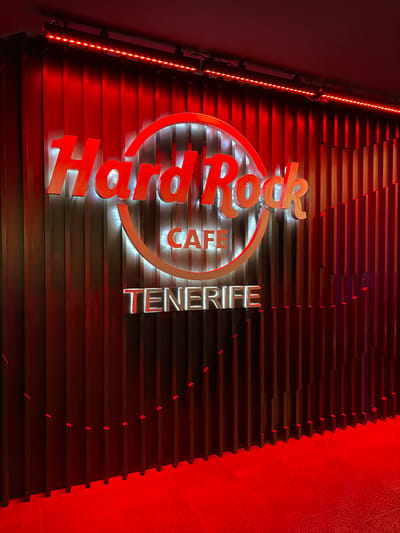The image shows the illuminated Hard Rock Cafe Tenerife sign on a wall composed of horizontal slats. The location is implied to be in Los Cristianos, Spain. The sign is brightly lit, with red and white colors. The lighting creates a strong contrast between light and shadow on the textured wall. The overall impression is one of a modern, stylish, and well-lit establishment.