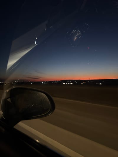 The image, captured from inside a car, shows a nighttime cityscape. The sky transitions from dark blue to a vibrant orange near the horizon. A line of lights, likely from buildings or streetlights, marks the edge of the city. Inside the car, the dashboard is visible, though details are obscured by the darkness. The car's side mirror is prominently featured, reflecting the surrounding environment. The interior of the car is mostly dark, with faint light illuminating some surfaces. Based on the location provided, Bol'shaya Mos', Russia, the cityscape likely represents the urban sprawl of the Moscow metropolitan area.