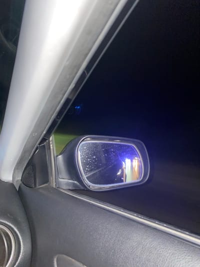 The image is taken from inside a vehicle, looking out the driver's side window at night. A side-view mirror is visible, reflecting lights that appear to be from another vehicle, possibly with flashing blue lights, suggesting a law enforcement presence. The interior of the car, including the door panel and speaker grille, is visible in the foreground. The background is dark, indicating nighttime. There are specks of what appear to be water droplets on the mirror. The location in the city of Yasen, Bulgaria is not determinable from the visual information provided.