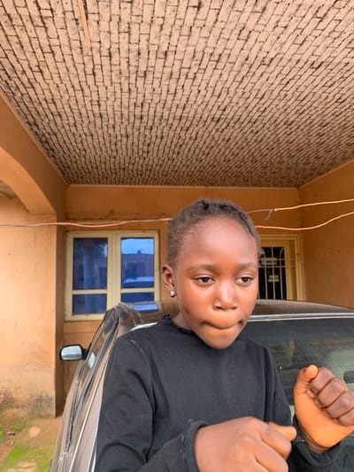 A young Black individual is the main subject, positioned in the foreground leaning out of a car. The individual wears a black long-sleeved shirt and is looking towards the camera with a slight smile. Their hands are raised, and they appear to be gesturing or posing. The background consists of a brick wall and a building with a window that has blue panes. The scene is outdoors, possibly in a residential area, with wires visible overhead. The lighting suggests it could be daytime. There is no visible text in the image. The image is oriented vertically.