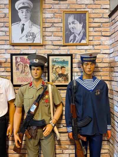 The image shows a brick wall adorned with historical and military-themed items. Two mannequins are dressed in Vietnamese military uniforms, each holding a rifle. One wears a navy uniform, and the other a khaki green army uniform with medals. Framed portraits of historical figures hang beside them, along with framed propaganda posters featuring sailors and soldiers. One poster reads, "Hai Quan Viet Nam," while another says, "Quan Doi Ta."