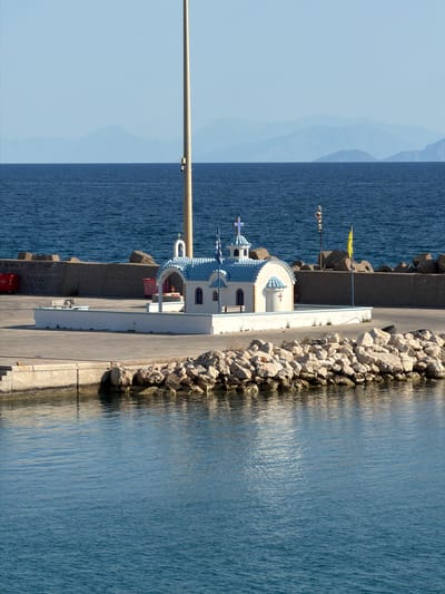 This image depicts a small, white church with a blue tiled roof, featuring a cross on its dome, situated on a paved pier next to a body of water. The church is decorated with blue trim and has a blue flag flying beside it. The pier is lined with rocks and a concrete barrier, with the sea stretching out to the horizon under a clear, pale blue sky. In the background, faint mountain ranges are visible. The scene is part of the Municipal Unit of Kastro - Kyllini, Greece.