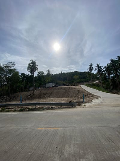The image depicts a concrete road in Baan Wang Ta Kien, Thailand. The road curves along the edge of a hillside densely covered with trees, including palm trees. A rough, dirt shoulder separates the road from the hillside. A metal guardrail runs along the roadside. The sky is visible above the trees and is partly cloudy with the sun shining through. There are no visible people.