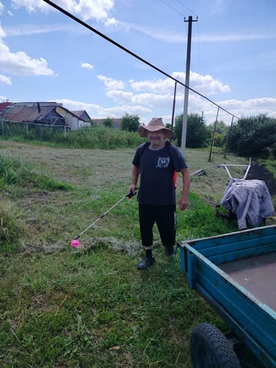 In an outdoor scene in Блохино, Russia, a man wearing a wide-brimmed hat and a dark t-shirt stands in a grassy yard. He is holding a long, thin pole with a pink object at its end, possibly a weed wacker or a similar gardening tool. The man is smiling and appears to be engaged in yard work.
Behind him, a power pole stands tall with wires extending across the frame. To his right, a garden tiller covered with a light-colored cloth is visible, along with a blue metal trailer. The background features houses with weathered roofs and dense green trees, suggesting a rural or semi-rural environment. The sky is bright blue with scattered white clouds, indicating a pleasant, possibly midday, time of day and fair weather. The overall impression is of a man working outdoors on a sunny day in a rustic setting.