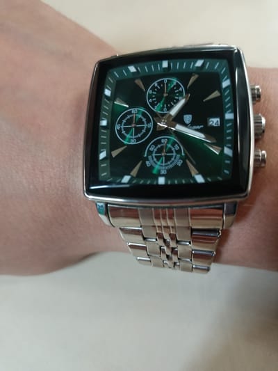 A close-up shot shows a person's wrist wearing a silver-banded, square-faced watch with a green dial. The watch has multiple subdials and a date window showing "24". The background is a plain, light-colored surface. There is no visible information in the image that would indicate the location in Mikhnevo, Russia.