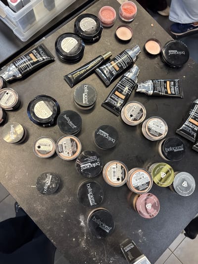 The image displays a cluttered array of cosmetic products, primarily from the Bellapierre brand, spread across a dark, matte surface, likely a table or counter, in Herentals, Belgium. Numerous black squeeze tubes, identified as Bellapierre BB Cream (SPF 20 Derma Renew) in shades such as 'Dark,' 'Light,' and 'Medium,' are visible, some appearing used. One smaller black tube of Bellapierre HD Moisturizing Primer is also present. Several black compacts bearing the Bellapierre logo are scattered, with some open to reveal internal labels or product.

A significant quantity of small, clear plastic jars with black or gold screw-on caps, containing various loose mineral powders (e.g., 'HD Finishing Powder,' 'Mineral Foundation,' 'Banana Setting Powder,' 'Honey'), are dispersed throughout the scene. Several jars have visible barcodes and product details on their labels. Two small open jars contain pink and orange-toned cream or blush products, and two detached black caps lie nearby. The dark surface is extensively covered with fine, white powder residue, indicating active use or spillage of the products.

In the upper left portion of the image, a clear plastic organizer bin holds additional cosmetic items. In the upper right background, the lower legs and feet of a person wearing light-colored pants and dark blue sneakers are partially visible. Another person's lower leg, clad in dark trousers, is visible in the lower left background. A black-handled brush or applicator is partially visible in the bottom right corner.