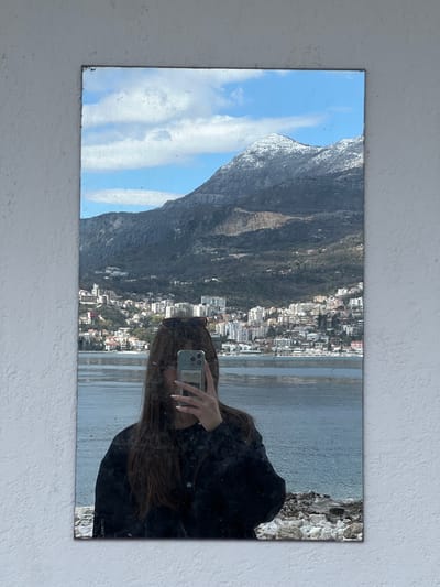 The image shows a rectangular mirror mounted on a white wall. The reflection in the mirror depicts a mountainous landscape with a city nestled along the coast. The city appears to be Provodina, Montenegro, indicated by the clustered buildings along the bay and the prominent mountain rising behind it. The right side of the mirror reflects a person taking a photo with a smartphone. The person is wearing a black jacket and has long brown hair. The background behind the person is not visible, as the reflection is focused on the cityscape. The sky is blue with clouds, and the sea is calm.